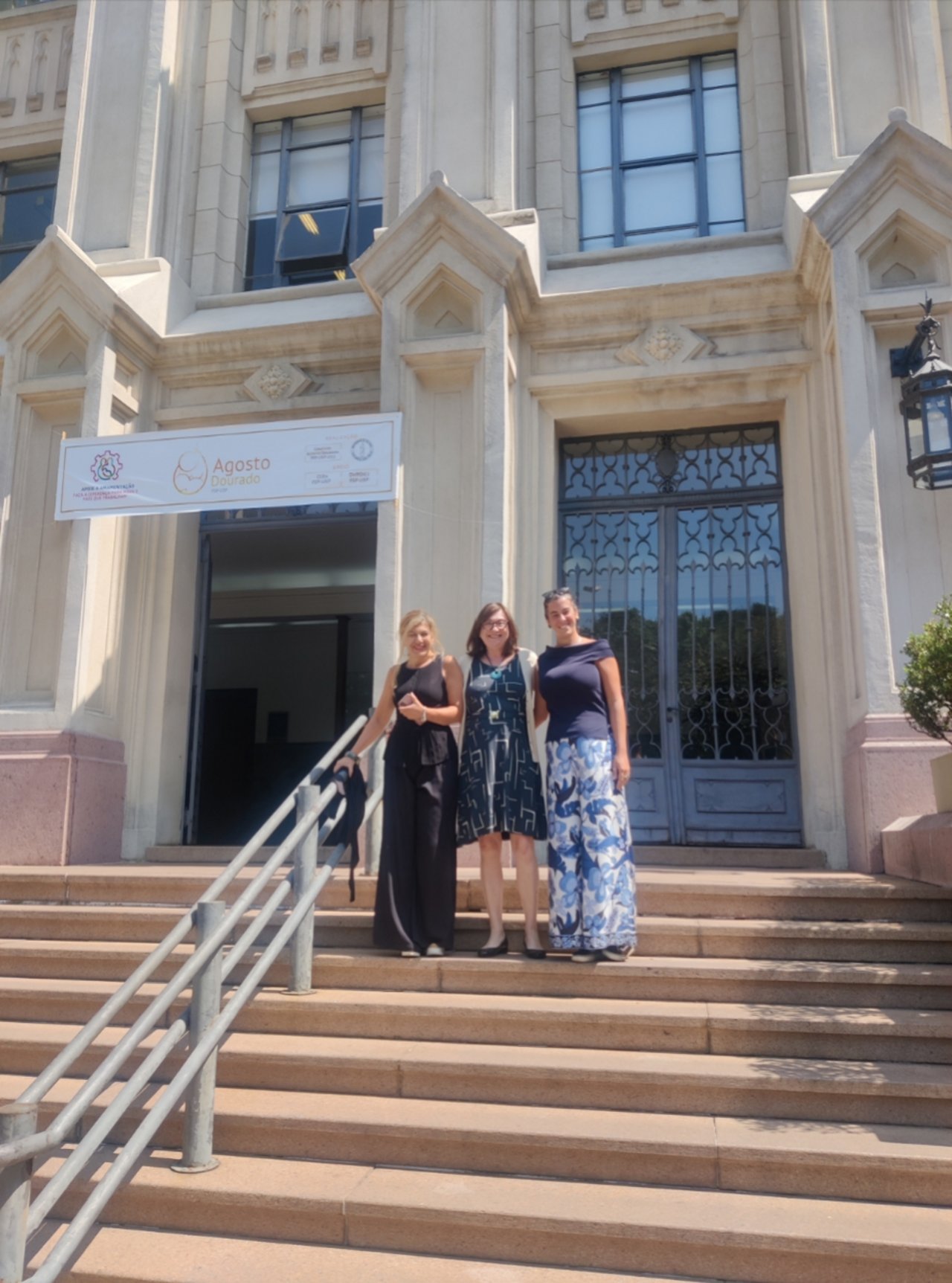 Tre persone posano sorridenti sui gradini davanti all’ingresso monumentale dell’Università di San Paolo, in Brasile, con grandi finestre, portone in ferro e una locandina appesa sulla facciata